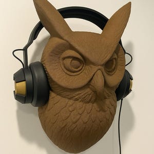 May include: A brown owl head wall mount with black headphones. The owl has detailed feather textures and is wearing black headphones with gold accents. The headphones' cord hangs down. The owl is mounted on a white wall.