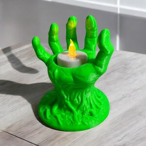 May include: A bright green, hand-shaped candle holder with a textured surface. The fingers cradle a white candle with a flickering yellow LED flame. The base is round and the overall design is reminiscent of a zombie hand, perfect for Halloween decor.