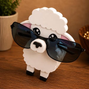 May include: A white sheep-shaped sunglasses holder with black sunglasses. The holder has a fluffy cloud-like top and black feet. The sheep's face is white with black eyes and a black nose. The sunglasses have a black frame and dark blue lenses.