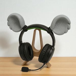 May include: Black over-ear headphones with a microphone, resting on a wooden stand. The headphones have gray, wing-shaped attachments. The stand is made of light-colored wood and has a simple, A-frame design. The background is a plain white wall.