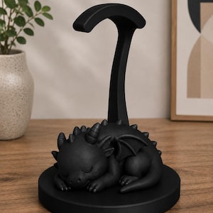 May include: A black dragon-shaped headphone stand. The dragon is sleeping on a round base, with a tall, curved support for headphones. The stand is made of a matte black material and is designed to hold headphones.