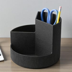 K&ouml;nnte beinhalten: Ein schwarzer Schreibtisch-Organizer mit drei F&auml;chern. Der Organizer hat eine strukturierte Oberfl&auml;che und enth&auml;lt blaue Scheren und einen gelben Bleistift. Das Design umfasst einen runden Sockel und einen hohen, abgewinkelten Abschnitt.