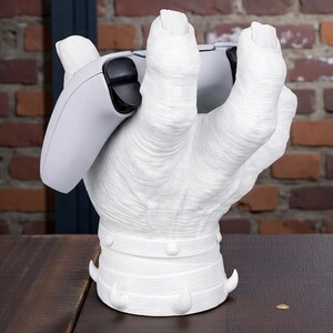 May include: A white, hand-shaped gaming controller stand. The stand is holding a white and black gaming controller. The base of the stand has decorative elements. The background features a brick wall.