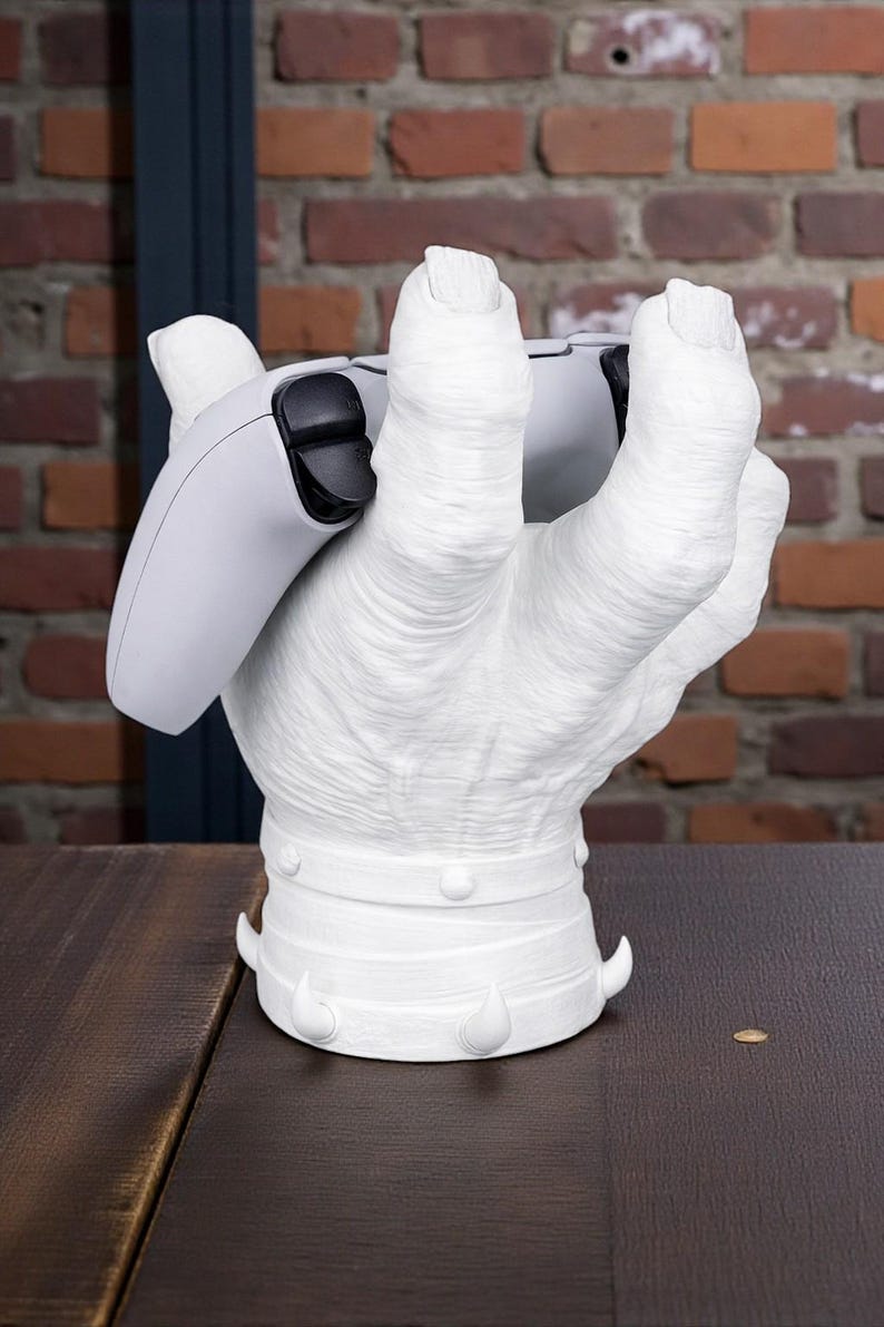 May include: A white, hand-shaped gaming controller stand. The stand is holding a white and black gaming controller. The base of the stand has decorative elements. The background features a brick wall.