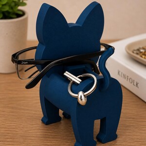 May include: A navy blue cat-shaped organizer holds eyeglasses and a set of keys. The wooden cat has a tail and is standing on a wooden surface. The eyeglasses are black-framed.