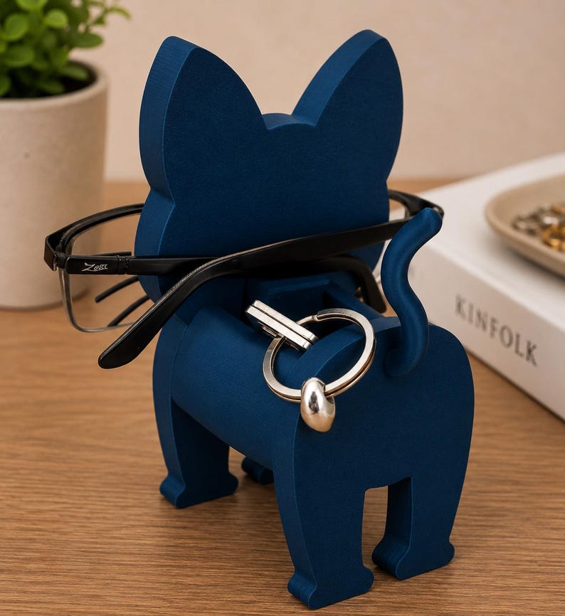 May include: A navy blue cat-shaped organizer holds eyeglasses and a set of keys. The wooden cat has a tail and is standing on a wooden surface. The eyeglasses are black-framed.