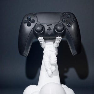 Op de afbeelding: Een zwarte gamecontroller wordt omhoog gehouden door een wit astronautenbeeldje. De controller heeft meerdere knoppen en richtingspads. De astronaut wordt ondersteund door een witte wolk-vormige basis.
