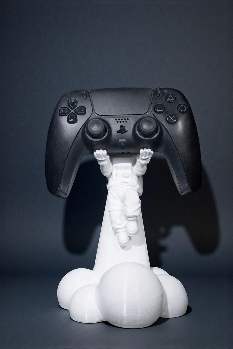 Op de afbeelding: Een zwarte gamecontroller wordt omhoog gehouden door een wit astronautenbeeldje. De controller heeft meerdere knoppen en richtingspads. De astronaut wordt ondersteund door een witte wolk-vormige basis.