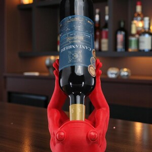 May include: A red wine bottle holder shaped like hands supporting a wine bottle. The bottle has a blue label with white text. The holder is on a wooden surface, with a bar and shelves of bottles in the background.