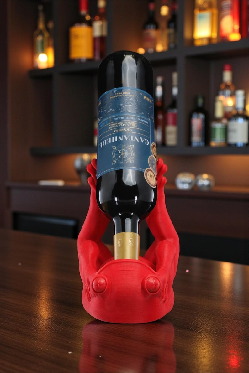 May include: A red wine bottle holder shaped like hands supporting a wine bottle. The bottle has a blue label with white text. The holder is on a wooden surface, with a bar and shelves of bottles in the background.