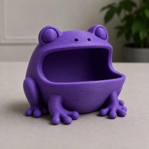 May include: A purple frog-shaped container with an open mouth, perfect for holding small items. The container has a textured surface and is designed with large eyes and detailed feet. The frog is a vibrant shade of purple.