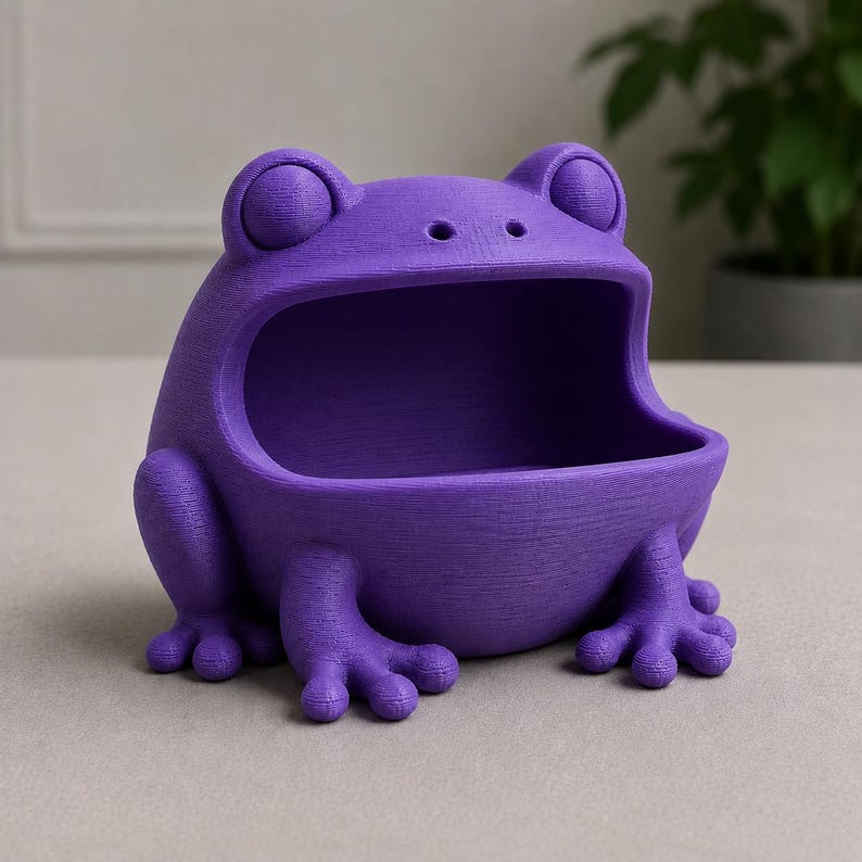 May include: A purple frog-shaped container with an open mouth, perfect for holding small items. The container has a textured surface and is designed with large eyes and detailed feet. The frog is a vibrant shade of purple.