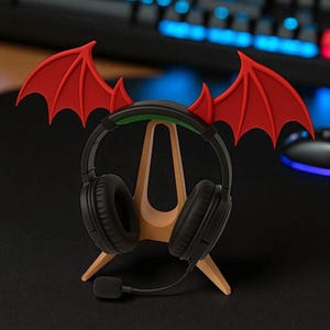 Op de afbeelding: Zwarte gaming-koptelefoon met microfoon op een houten standaard. De standaard is versierd met rode duivelsvleugels. De koptelefoon staat op een zwart oppervlak, met een toetsenbord op de achtergrond.
