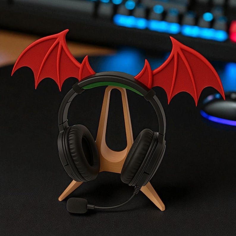 Op de afbeelding: Zwarte gaming-koptelefoon met microfoon op een houten standaard. De standaard is versierd met rode duivelsvleugels. De koptelefoon staat op een zwart oppervlak, met een toetsenbord op de achtergrond.