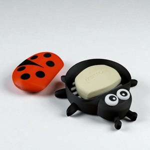 May include: A whimsical black ladybug-shaped soap dish with a matching orange lid. The dish holds a bar of soap with the text "MEN+CARE". The ladybug has large, cartoonish eyes and legs.