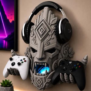 May include: A gray stone-textured wall mount shaped like a carved mask with two claw-like hands holding a white and a black gaming controller. The mask features a glowing blue light in its open mouth and supports a pair of black over-ear headphones.