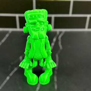 May include: A bright green 3D printed figurine of Frankenstein's monster. The figurine has a goofy expression and is standing with its arms outstretched.