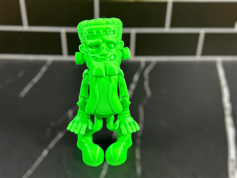 May include: A bright green 3D printed figurine of Frankenstein's monster. The figurine has a goofy expression and is standing with its arms outstretched.