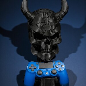 以下が含まれることがあります： 角のある黒い頭蓋骨型のゲーム機ホルダーと青いゲームコントローラー。 頭蓋骨は詳細でテクスチャ加工されたデザインです。 コントローラーは標準サイズです。