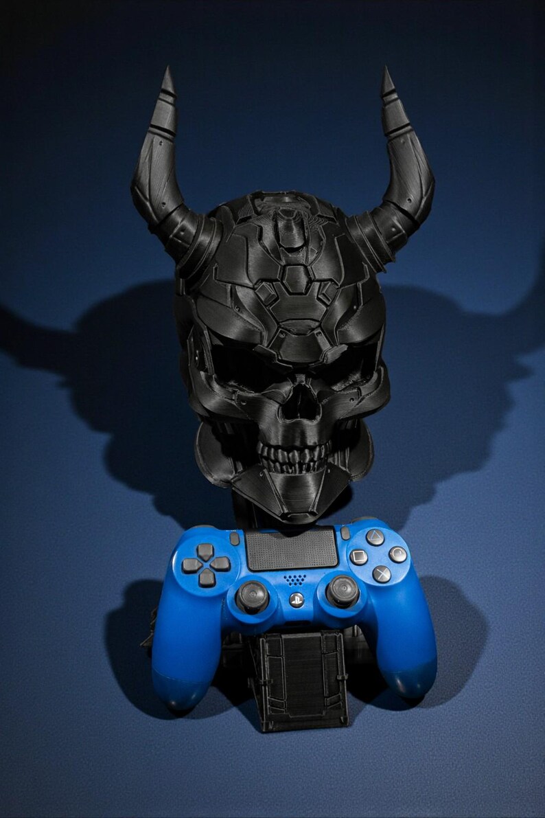 以下が含まれることがあります： 角のある黒い頭蓋骨型のゲーム機ホルダーと青いゲームコントローラー。 頭蓋骨は詳細でテクスチャ加工されたデザインです。 コントローラーは標準サイズです。