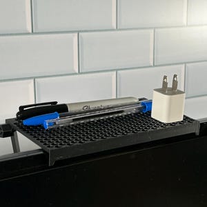 May include: A black plastic desk organizer with a grid pattern, holding two pens and a white phone charger. The organizer is attached to the top of a black computer monitor.