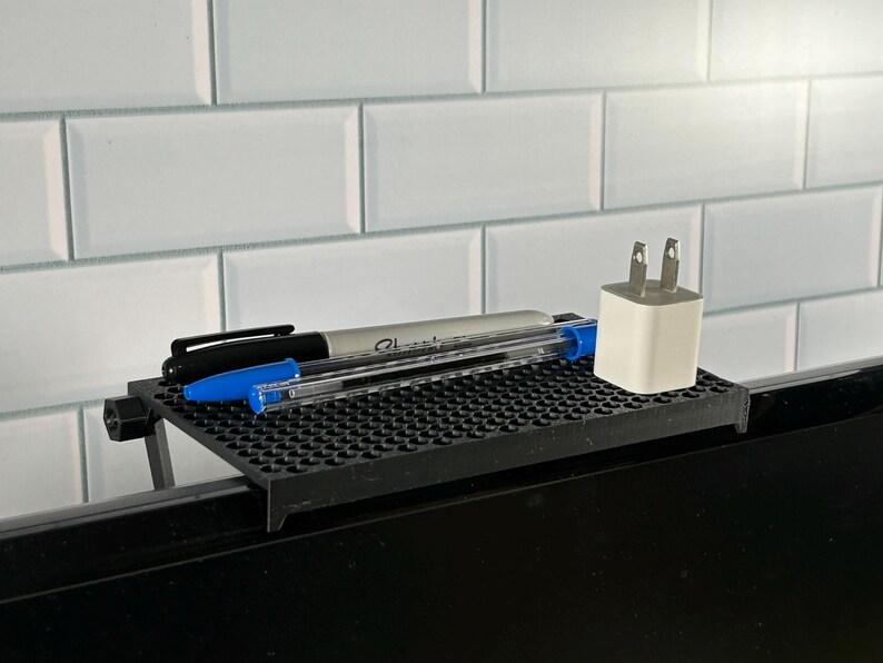 May include: A black plastic desk organizer with a grid pattern, holding two pens and a white phone charger. The organizer is attached to the top of a black computer monitor.