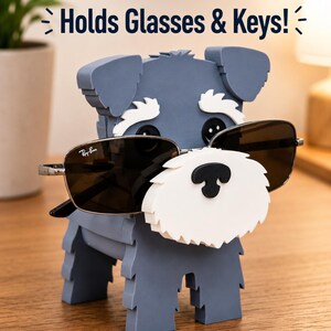 May include: A gray dog-shaped glasses holder with a white beard and black nose, holding sunglasses. The text "Holds Glasses & Keys!" is above the dog. The sunglasses are Ray-Ban brand.