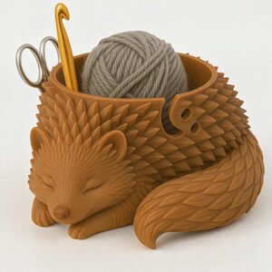 May include: A brown hedgehog-shaped yarn bowl with a ball of gray yarn inside. A gold crochet hook and silver scissors are placed in the bowl. The hedgehog is sleeping with its eyes closed.