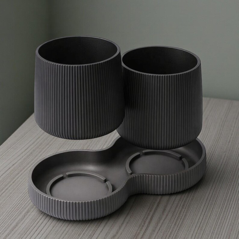 May include: A dark gray bathroom organizer set. The set includes two cylindrical cups and a tray with two circular compartments. The cups and tray have vertical ribbed detailing. The set is designed to hold toothbrushes and other bathroom essentials.