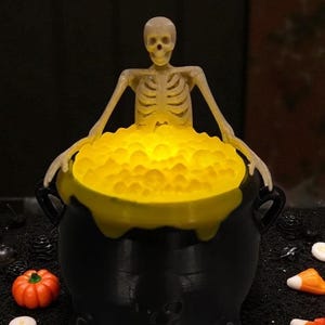 May include: A Halloween-themed decoration featuring a black cauldron filled with glowing yellow liquid. A plastic skeleton figure is emerging from the cauldron. A small orange pumpkin and candy corn are in the foreground.