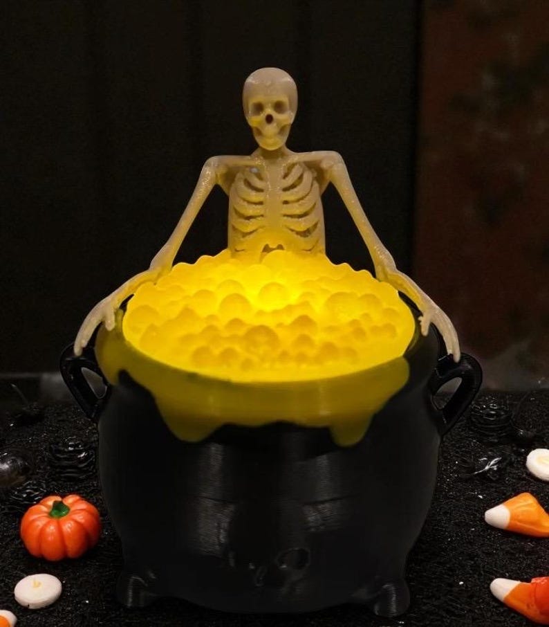 May include: A Halloween-themed decoration featuring a black cauldron filled with glowing yellow liquid. A plastic skeleton figure is emerging from the cauldron. A small orange pumpkin and candy corn are in the foreground.