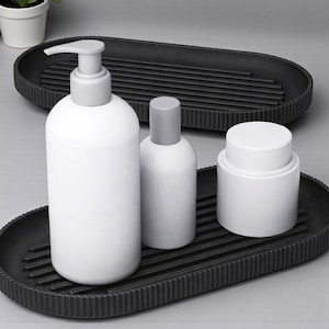 May include: A set of white cosmetic bottles and jars with gray lids on two black oval trays. The trays have a ribbed design and are placed on a light gray surface. A small potted plant is in the background.