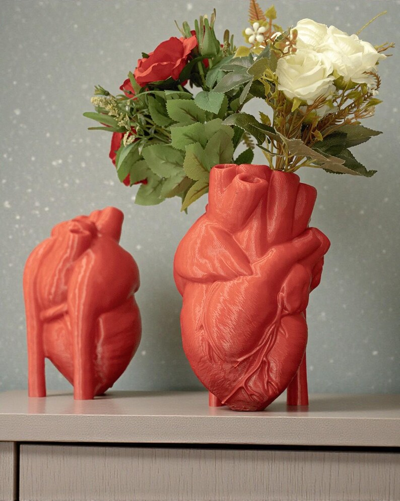 May include: Two red heart-shaped vases, one holding a bouquet of red and white roses with green leaves. The vases are on a gray surface against a speckled blue background. The vases are detailed with anatomical features.