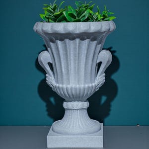 K&ouml;nnte beinhalten: Eine graue, verzierte Urne mit geriffeltem Design und zwei geschwungenen Griffen. Die Urne ist mit leuchtend gr&uuml;nen Kunstpflanzen gef&uuml;llt. Die Urne steht auf einem quadratischen Sockel, und der Hintergrund ist eine t&uuml;rkisfarbene Wand.