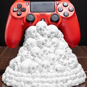 May include: A red video game controller rests above a white sculpture of stacked skulls. The controller has black buttons and directional pads. The sculpture is on a wooden surface.