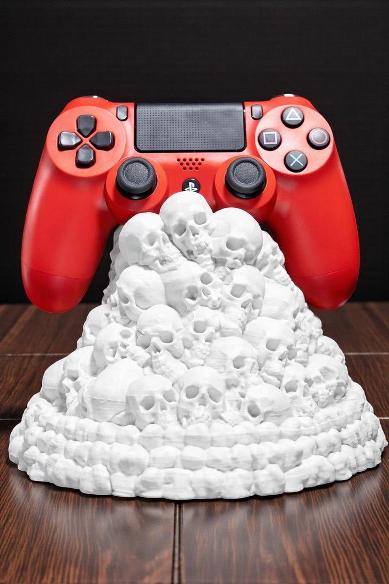 May include: A red video game controller rests above a white sculpture of stacked skulls. The controller has black buttons and directional pads. The sculpture is on a wooden surface.