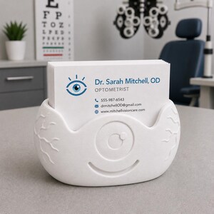 May include: A white, eye-shaped business card holder with a stack of white cards. The cards display the name "Dr. Sarah Mitchell, OD, Optometrist" and contact information. The holder has a textured surface and a smiling eye design.