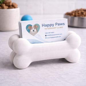 K&ouml;nnte beinhalten: Ein wei&szlig;er, knochenf&ouml;rmiger Visitenkartenhalter. Der Halter ist mit Karten gef&uuml;llt, auf denen "Happy Paws Veterinary Clinic" steht. Die Karten zeigen einen Cartoon-Hund in einem Herzen. Kontaktinformationen sind ebenfalls auf den Karten sichtbar.