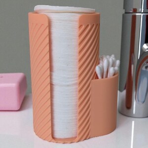 May include: A peach-colored, cylindrical bathroom organizer with a textured, diagonal stripe design. It holds a stack of white cotton pads and a small section for cotton swabs. A pink soap dish and a chrome bottle are in the background.