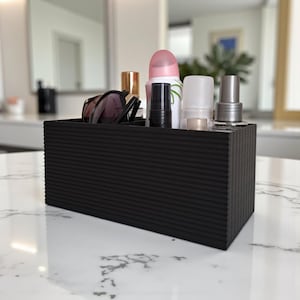 May include: A black, rectangular organizer with a ribbed texture, holding various beauty products. The organizer contains sunglasses, lipstick, a spray bottle, and other cosmetic items. The container sits on a white marble surface.