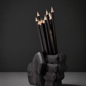 May include: A black pencil holder shaped like a clenched fist, holding several black pencils with sharpened tips. The pencils are arranged vertically, with their erasers and tips visible. The background is a dark gray.