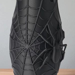 May include: Black vase with a spider web design and a large spider on the side. The vase has a unique shape and a textured surface, perfect for home decor or Halloween.