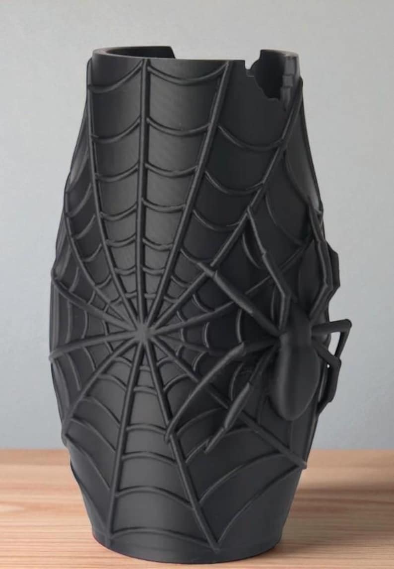 May include: Black vase with a spider web design and a large spider on the side. The vase has a unique shape and a textured surface, perfect for home decor or Halloween.