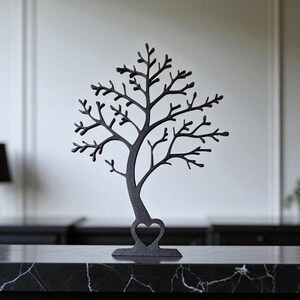 May include: A dark gray metal tree-shaped jewelry holder with a heart cutout at the base. The tree has many branches and is displayed on a black marble surface. The background is a neutral white wall.