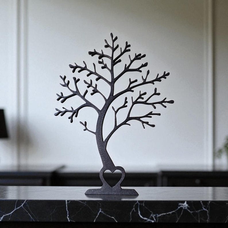 May include: A dark gray metal tree-shaped jewelry holder with a heart cutout at the base. The tree has many branches and is displayed on a black marble surface. The background is a neutral white wall.