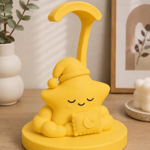 May include: A yellow star-shaped night light with a sleeping cap and a crescent moon-shaped pillow. The night light has a curved handle and is sitting on a round base. The star has closed eyes and a smiling mouth.