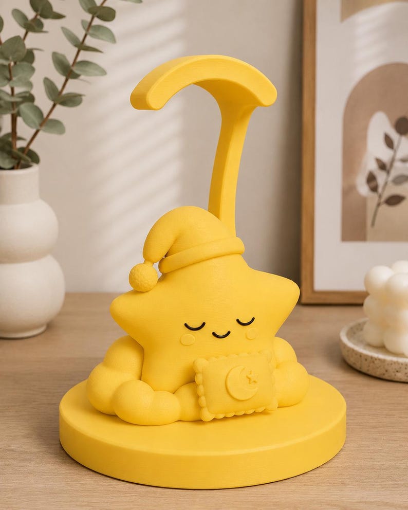 May include: A yellow star-shaped night light with a sleeping cap and a crescent moon-shaped pillow. The night light has a curved handle and is sitting on a round base. The star has closed eyes and a smiling mouth.