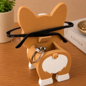 May include: A novelty corgi-shaped eyeglasses holder in tan and white. The holder has a slot for glasses and a key ring holder. Black eyeglasses are placed in the holder. The product is on a wooden surface.