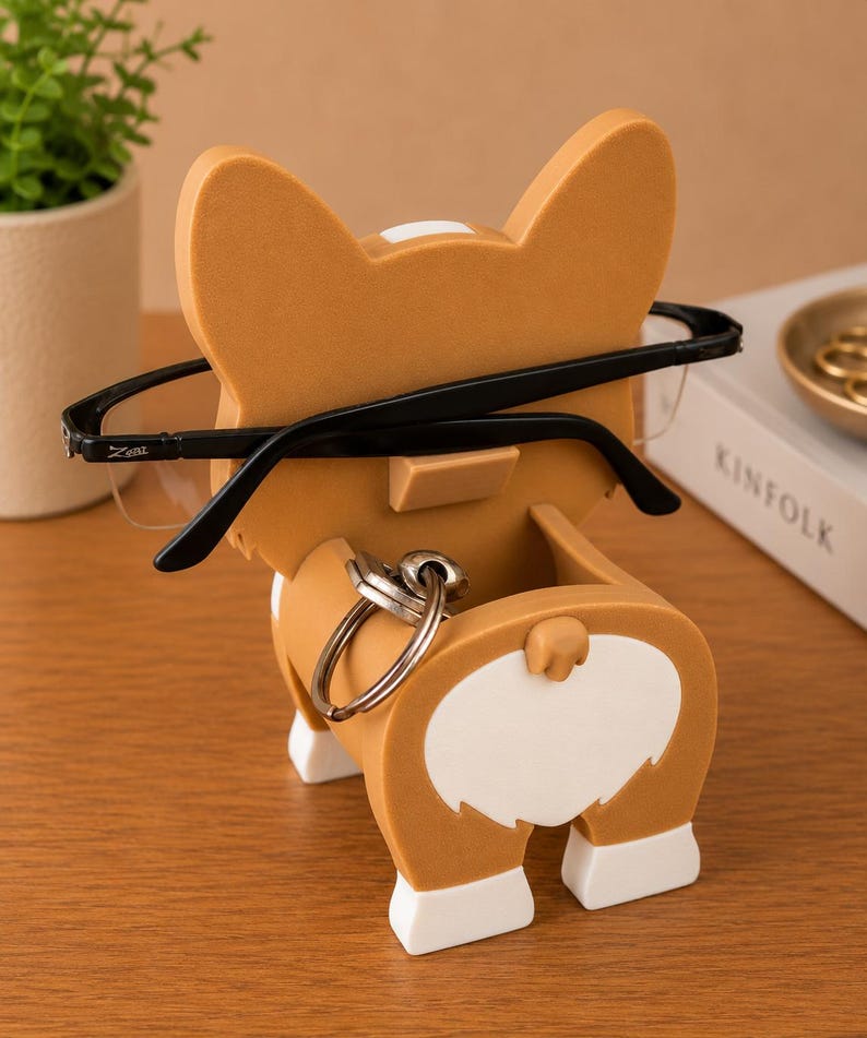 May include: A novelty corgi-shaped eyeglasses holder in tan and white. The holder has a slot for glasses and a key ring holder. Black eyeglasses are placed in the holder. The product is on a wooden surface.