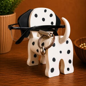 K&ouml;nnte beinhalten: Ein wei&szlig;-schwarz gefleckter Brillen- und Schl&uuml;sselhalter in Form eines Dalmatiners. Der Halter hat einen Schlitz f&uuml;r Brillen und einen Ring f&uuml;r Schl&uuml;ssel. Der Hunde-Halter ist aus Holz gefertigt und hat schwarze Ohren und schwarze Flecken.